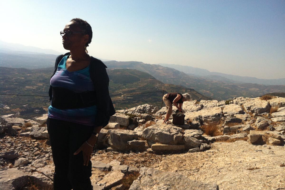 Two visitors stand on the rocky terrain at Juktas Peak Sanctuary, overlooking distant hills.