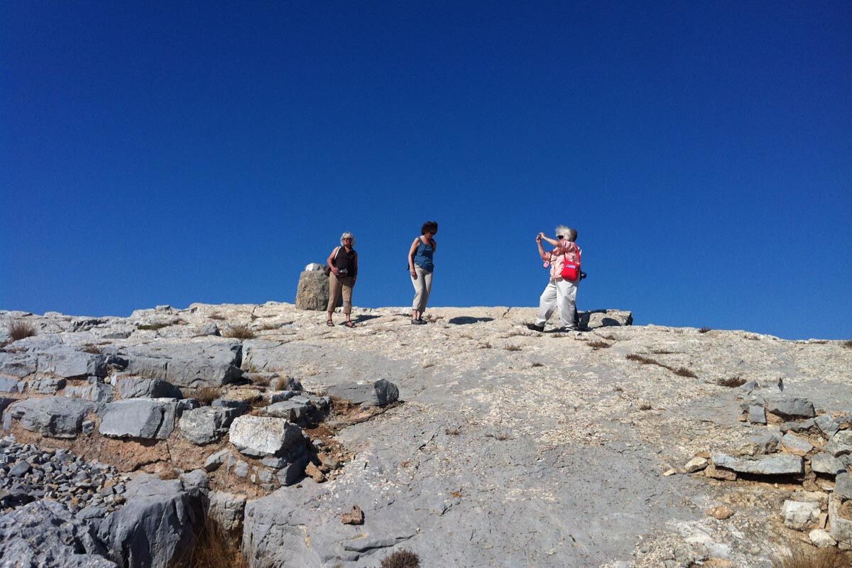 A small group of visitors stands on rocky ground at Juktas Peak Sanctuary under a clear blue sky.