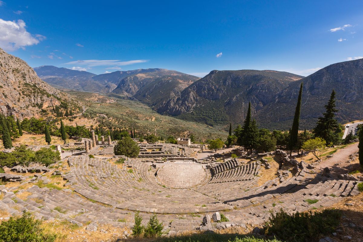 The ancient theater of Delphi, nestled on a hillside with stone seating overlooking a lush valley and surrounded by towering mountains under a clear blue sky.