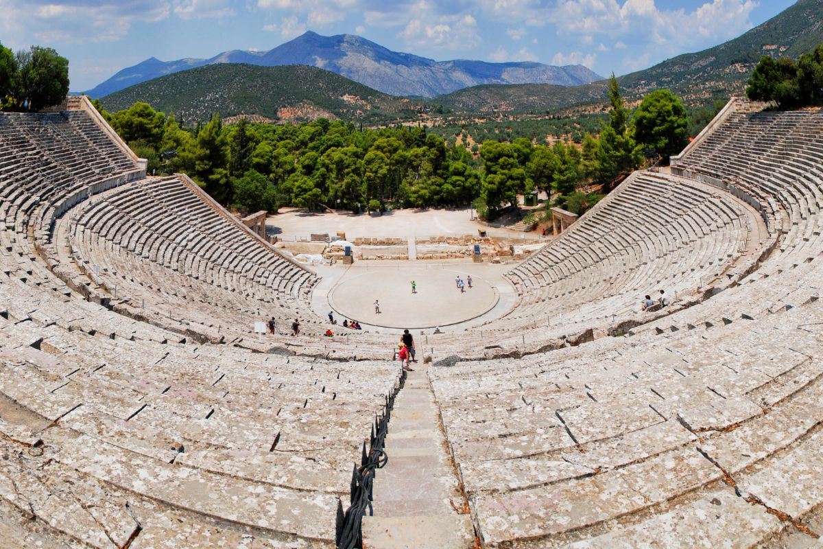 The ancient theater of Epidaurus, a well-preserved stone amphitheater with concentric seating, surrounded by lush greenery and distant mountains under a partly cloudy sky.