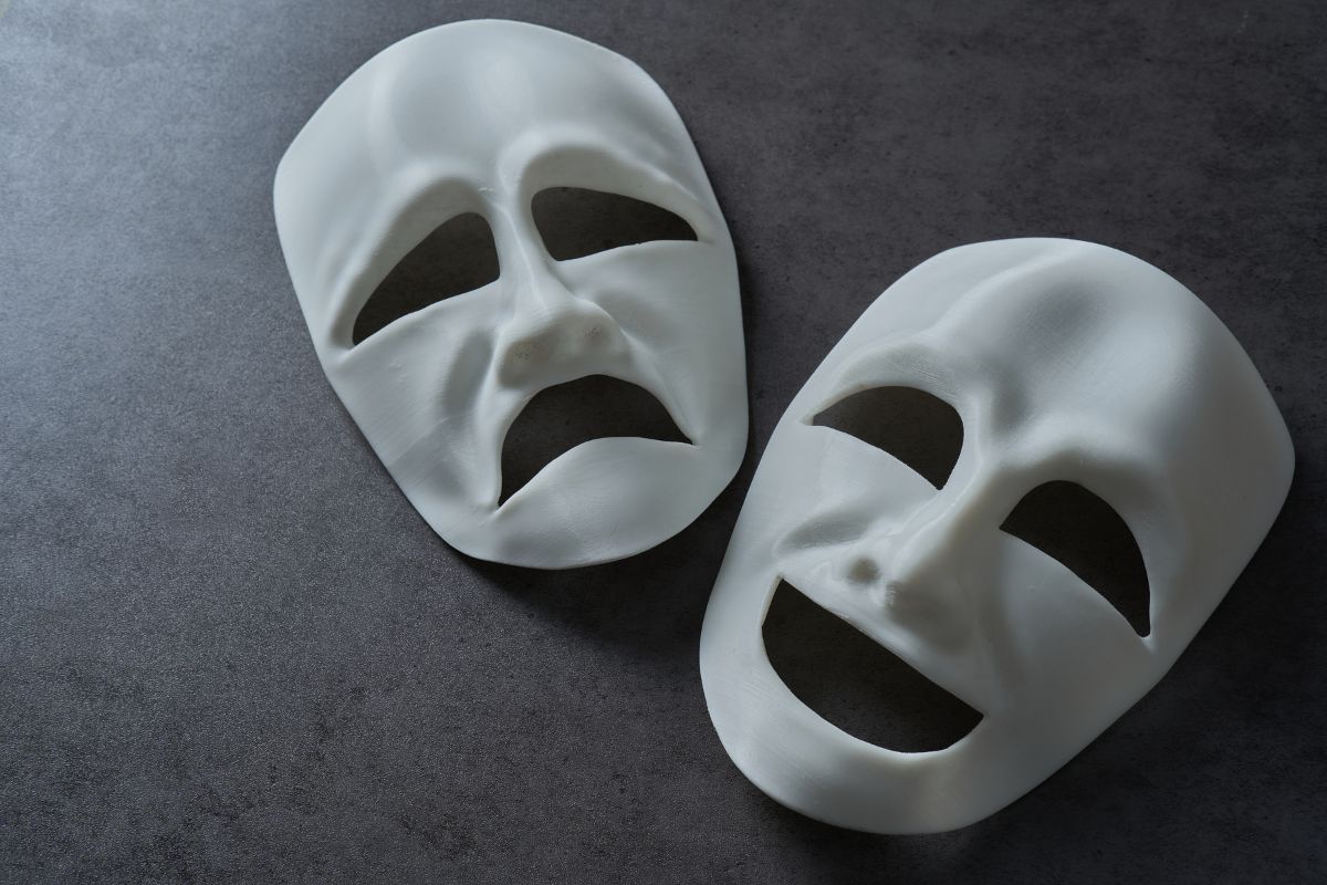 Two white ancient Greek theater masks on a dark surface, one depicting a joyful expression and the other a sorrowful one, symbolizing comedy and tragedy.