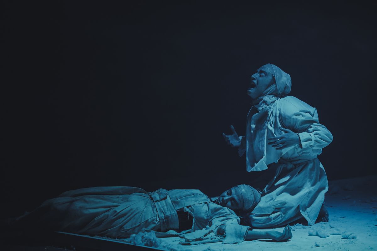 A dramatic scene from an ancient Greek tragedy, with one actor passionately lamenting while another lies motionless on the ground, lit by a moody blue light.