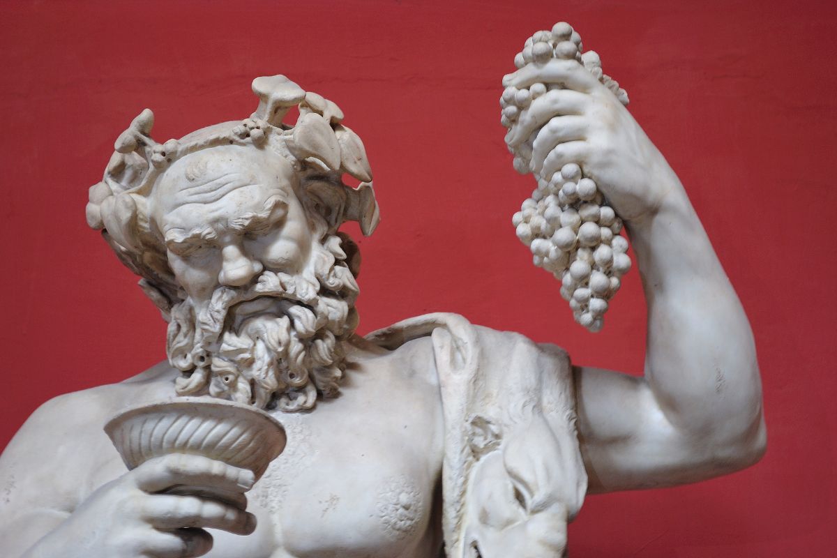 A marble statue of the Greek god Dionysus, holding a bunch of grapes in one hand and a cup in the other, symbolizing wine and festivity, set against a red background.