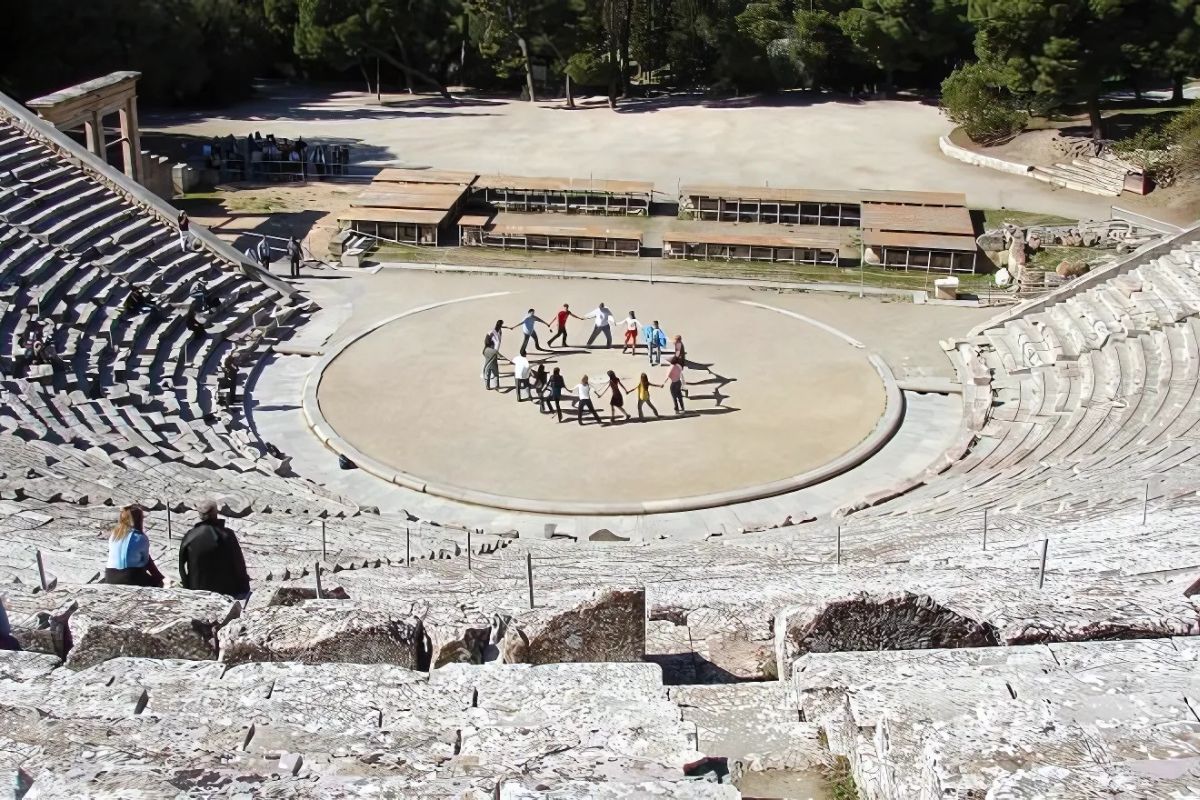 A group of people holding hands in a circle on the stage of an ancient Greek theater, participating in the Ellisos experience, surrounded by stone seating and greenery.