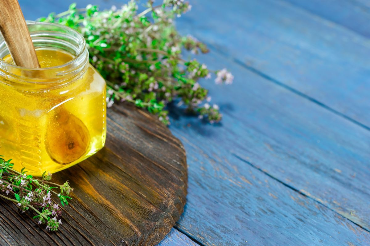 A jar of golden thyme honey with a wooden dipper, placed on a rustic wooden surface surrounded by sprigs of fresh thyme.