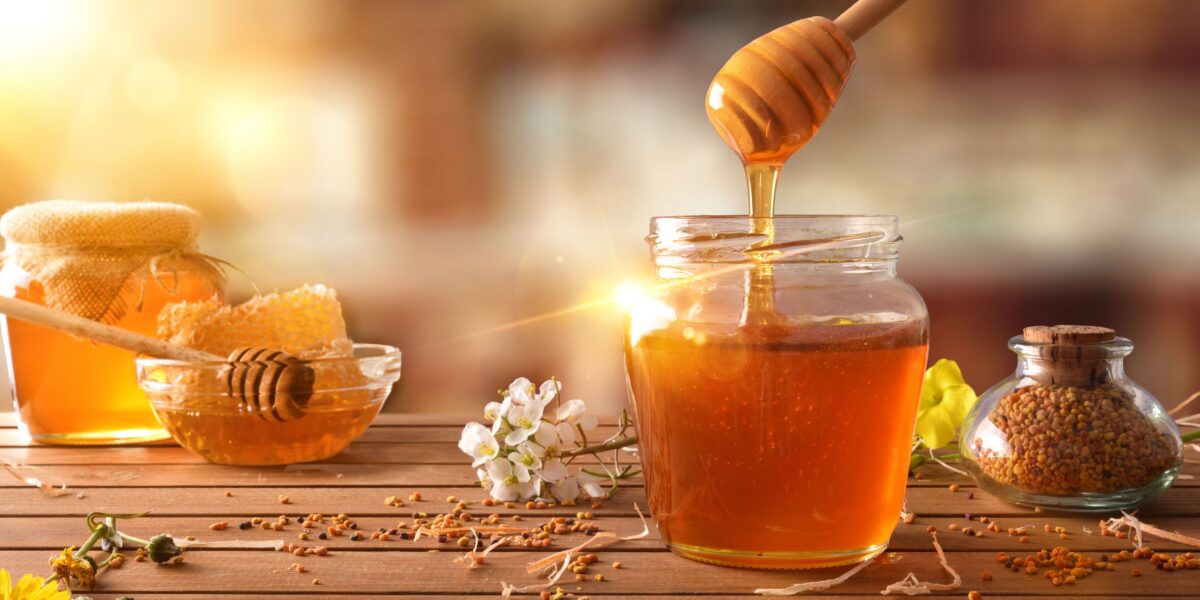 A jar of honey with a dipper, surrounded by honeycomb, flowers, and a small jar of pollen on a wooden table, with warm sunlight in the background.