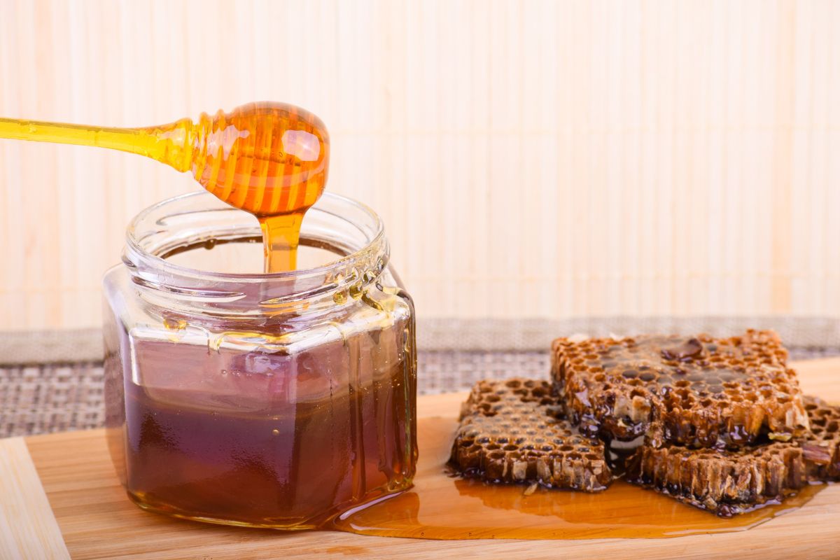 A jar of honey with a dipper resting on top, next to pieces of honeycomb on a wooden surface.