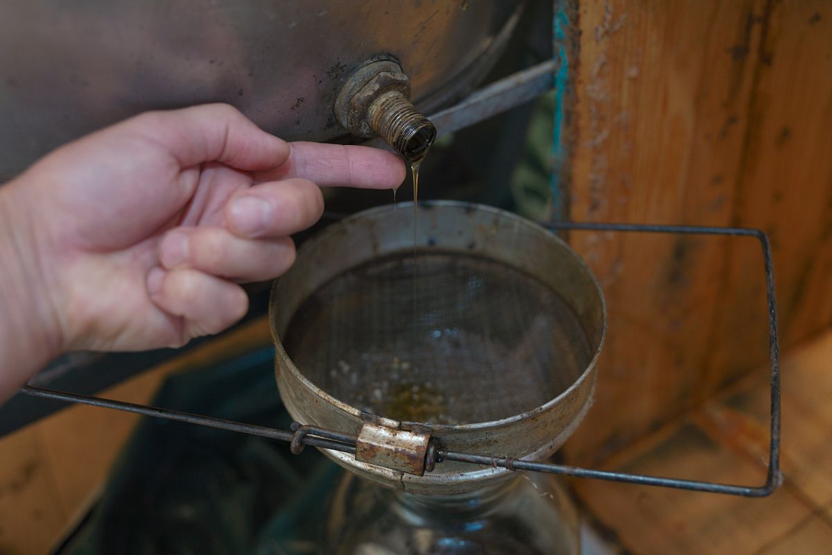 A person tasting honey directly from a metal spout, with a mesh filter and jar placed below.