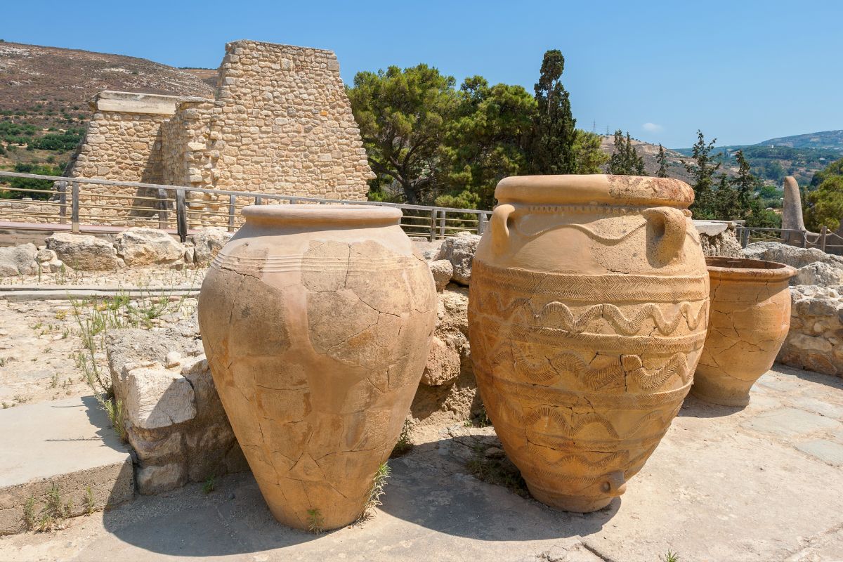 Large ancient clay storage jars (pithoi) at Knossos Palace, set against stone ruins and a background of trees and hills.