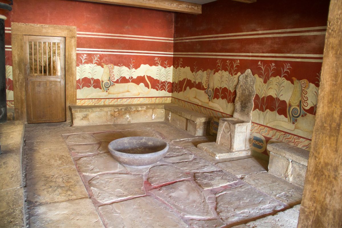 A room inside Knossos Palace featuring a stone throne, benches along the walls, a large bowl in the center, and red-painted walls with frescoes of mythical creatures.