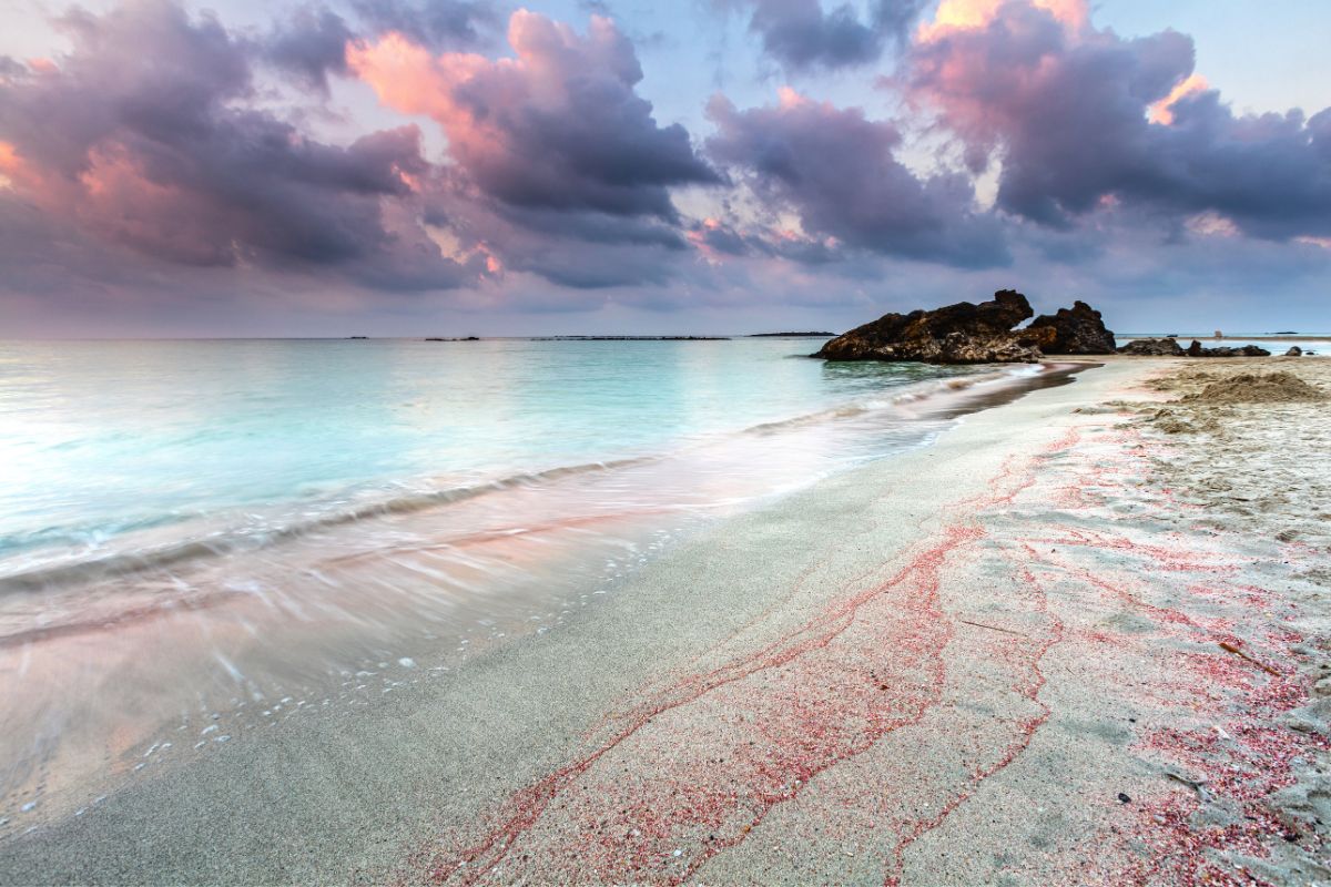 The serene shoreline of Elafonissi Beach in Crete, with pink-tinted sand, turquoise waters, and a dramatic, colorful sky. Weather in Crete in winter