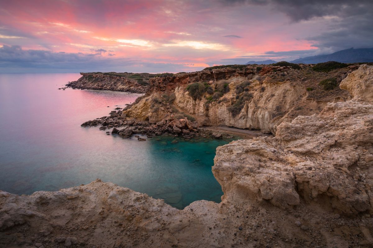 A serene coastal landscape in Crete at sunset, featuring rocky cliffs, turquoise waters, and a colorful sky.