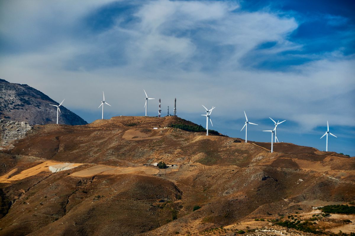 Wind turbines on a hill in Crete, set against a backdrop of rugged terrain and a partly cloudy sky. Weather in Crete in winter.