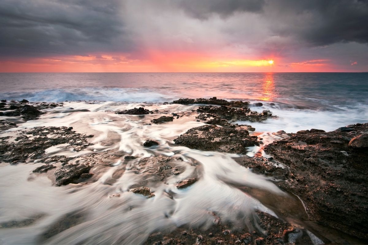 A rocky shoreline in Crete during sunset, with waves gently flowing over the rocks and vibrant colors in the sky.