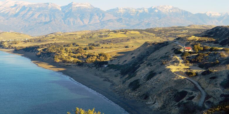 A scenic coastline in Crete with a sandy beach, rolling hills, and snow-capped mountains in the background.