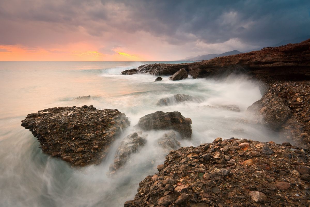 Rocky coastal scenery in Crete, with waves crashing against the rocks under a moody sky at sunset. Weather in Crete in winter