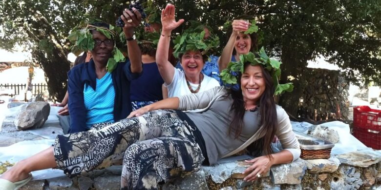 A group of smiling people wearing grapevine wreaths, raising their hands and celebrating during a wine-tasting experience in Crete.