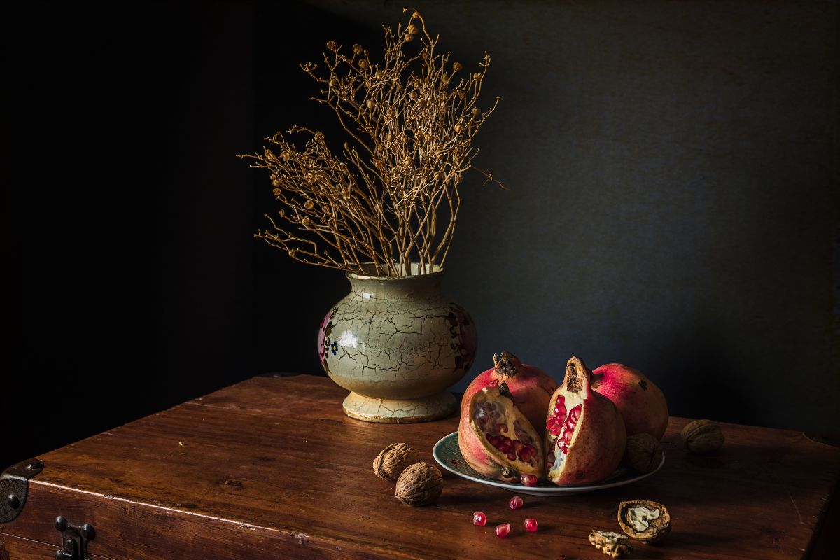 A still life featuring a ceramic vase with dried branches, pomegranates, and walnuts arranged on a wooden surface against a dark background.