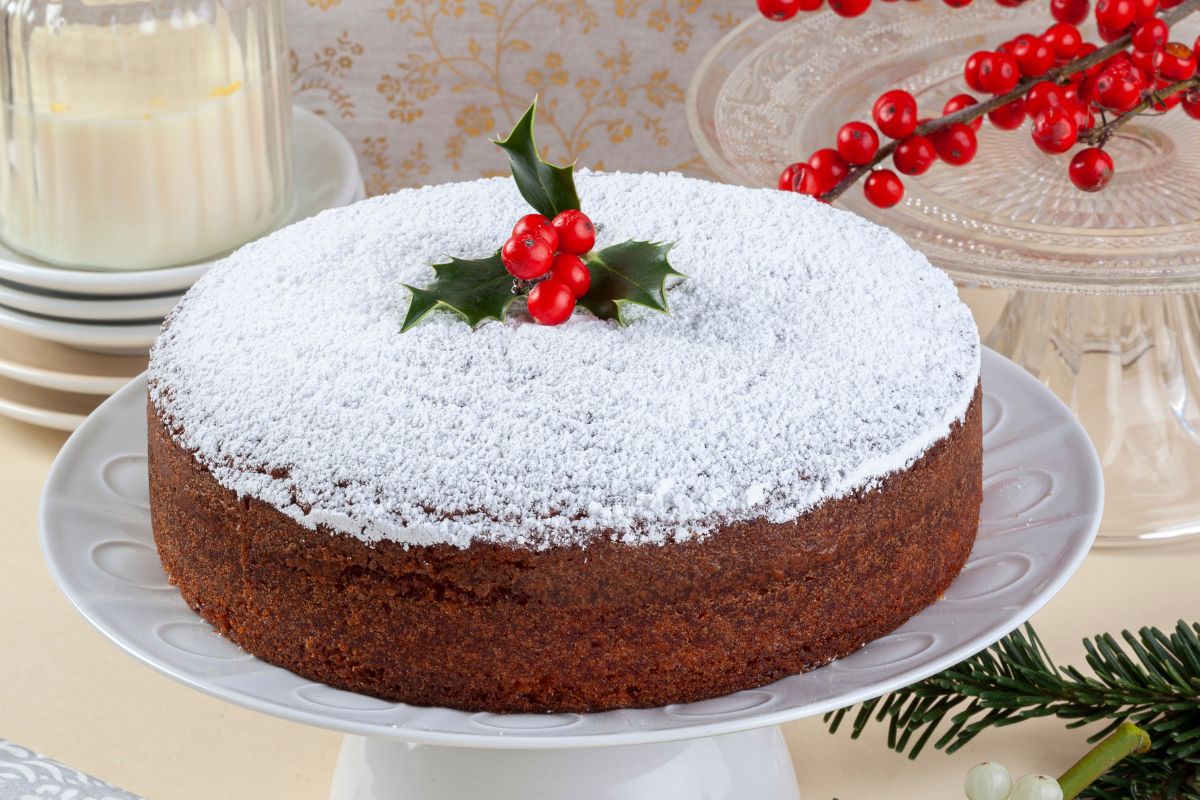 A traditional Vasilopita cake dusted with powdered sugar, topped with holly leaves and berries, displayed on a white cake stand with festive decor in the background.