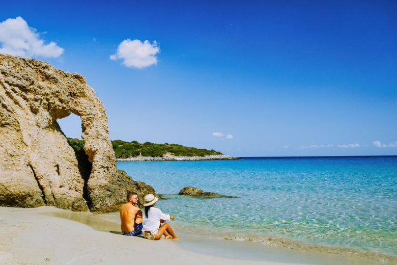 A couple sitting by a clear turquoise sea near a rocky arch formation on a sunny beach.