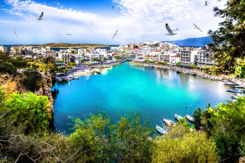 Agios Nikolaos Crete, a coastal town with vibrant blue water surrounded by buildings, trees, and boats, under a bright sky with a few flying birds.