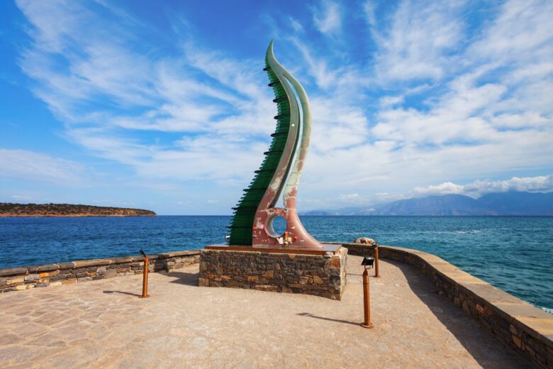 A unique monument in Agios Nikolaos Crete with a curved, green structure on a stone platform by the sea, under a partly cloudy sky.