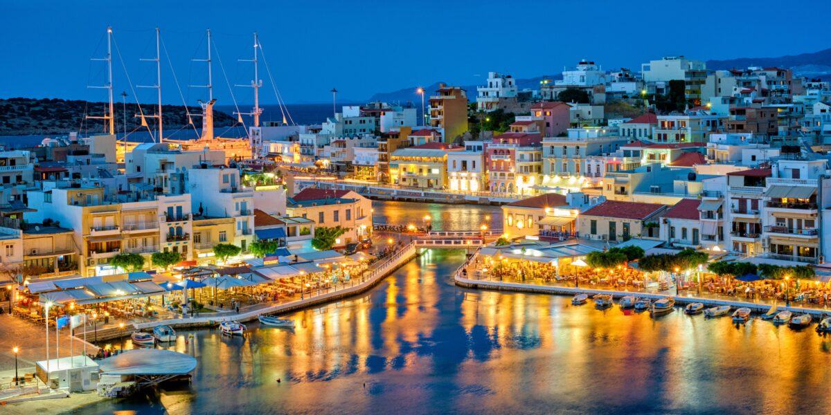 The vibrant coastal town of Agios Nikolaos at dusk with illuminated buildings reflecting on the water, featuring boats docked along the waterfront and a lively atmosphere.