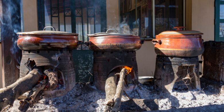 Three large clay pots sit on traditional wood-burning stoves, with logs fueling the fires beneath them. Smoke rises from the pots, indicating they are in use, likely for cooking.