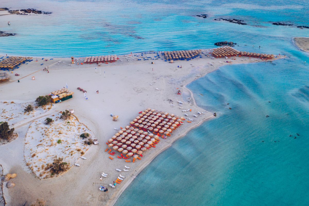 An aerial view of Elafonissi Beach in Crete, showing a sandy shore with organized sunbeds and umbrellas, surrounded by clear, turquoise waters.