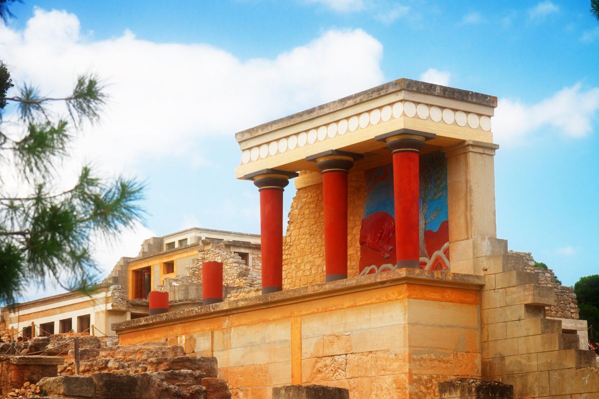 The image shows a section of the ancient Knossos Palace in Crete, featuring red columns and a mural with bright colors. The architecture is characterized by its historical Minoan style.