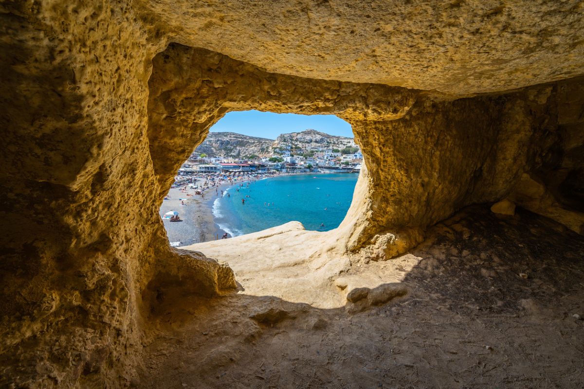 A view of a beach and seaside village through the opening of a cave in Matala, Crete, with clear blue water and people enjoying the beach.