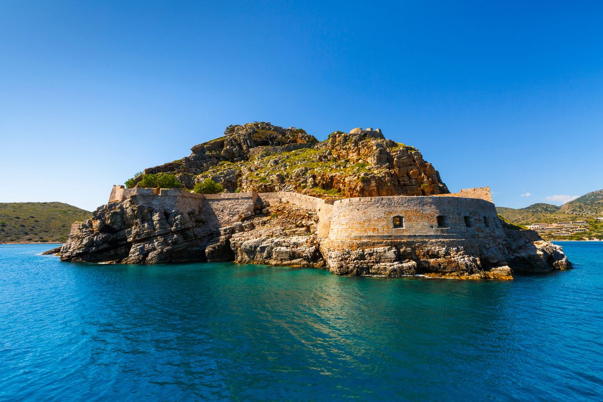 Spinalonga island in Crete with ancient stone fortifications surrounded by clear blue water under a bright blue sky.