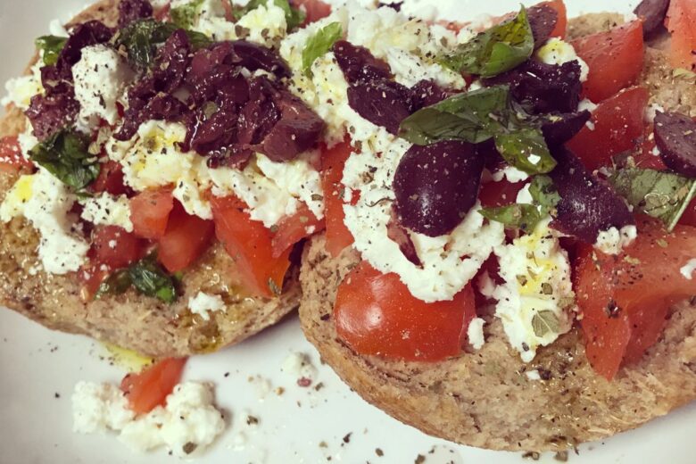 A plate of Cretan Dakos, featuring rusks topped with chopped tomatoes, crumbled cheese, olives, and fresh herbs.