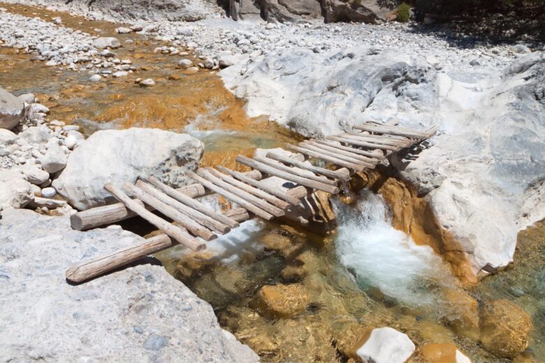 A small, rustic wooden bridge spans a rocky stream with clear, flowing water. The surrounding area is filled with smooth stones and boulders.