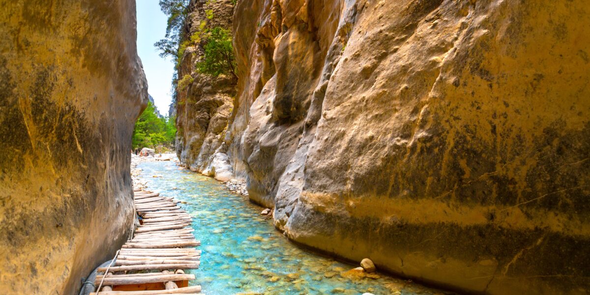 A narrow pathway with a wooden plank bridge runs alongside a turquoise stream, flanked by towering, steep rock walls in Samaria Gorge.