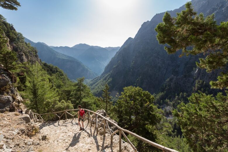 A hiker with a red backpack walks along a rocky trail with a wooden railing, surrounded by lush green trees and overlooking a valley with tall, rugged mountains.