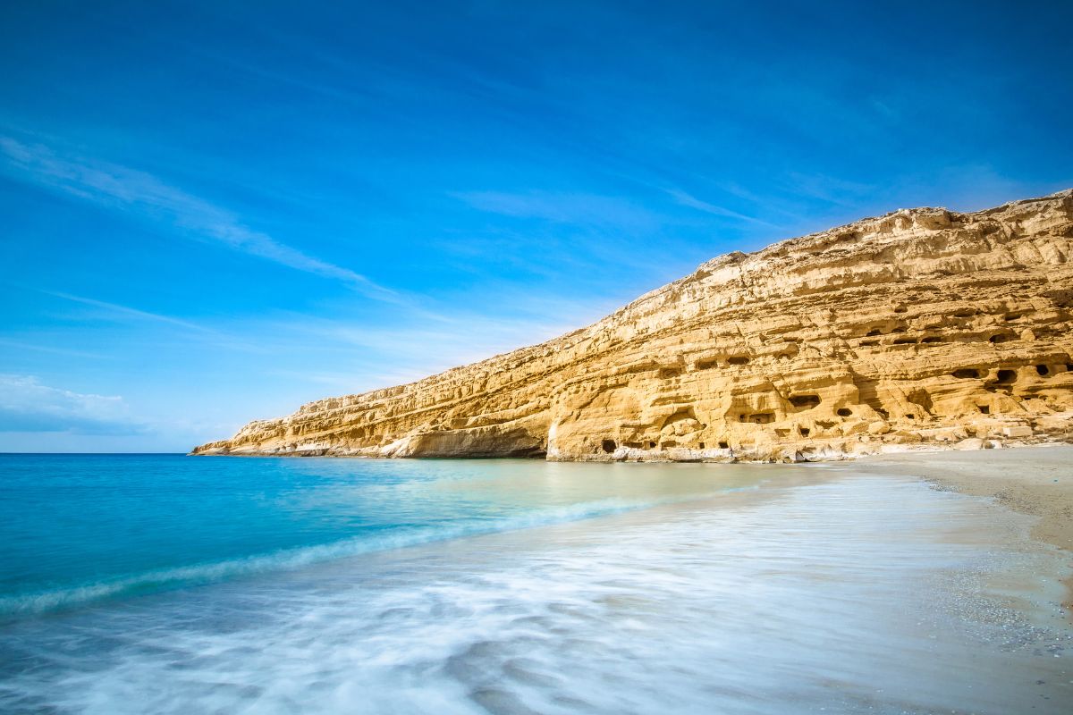 Matala Beach with its unique sandstone cliffs and calm turquoise waters under a clear blue sky. The shoreline shows gentle waves washing onto the sandy beach.