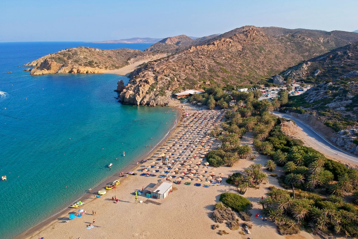 Aerial view of Vai Beach featuring golden sands, clear blue waters, and rows of umbrellas and sunbeds. The beach is surrounded by rocky hills and lush palm trees, with a parking area and road visible nearby.