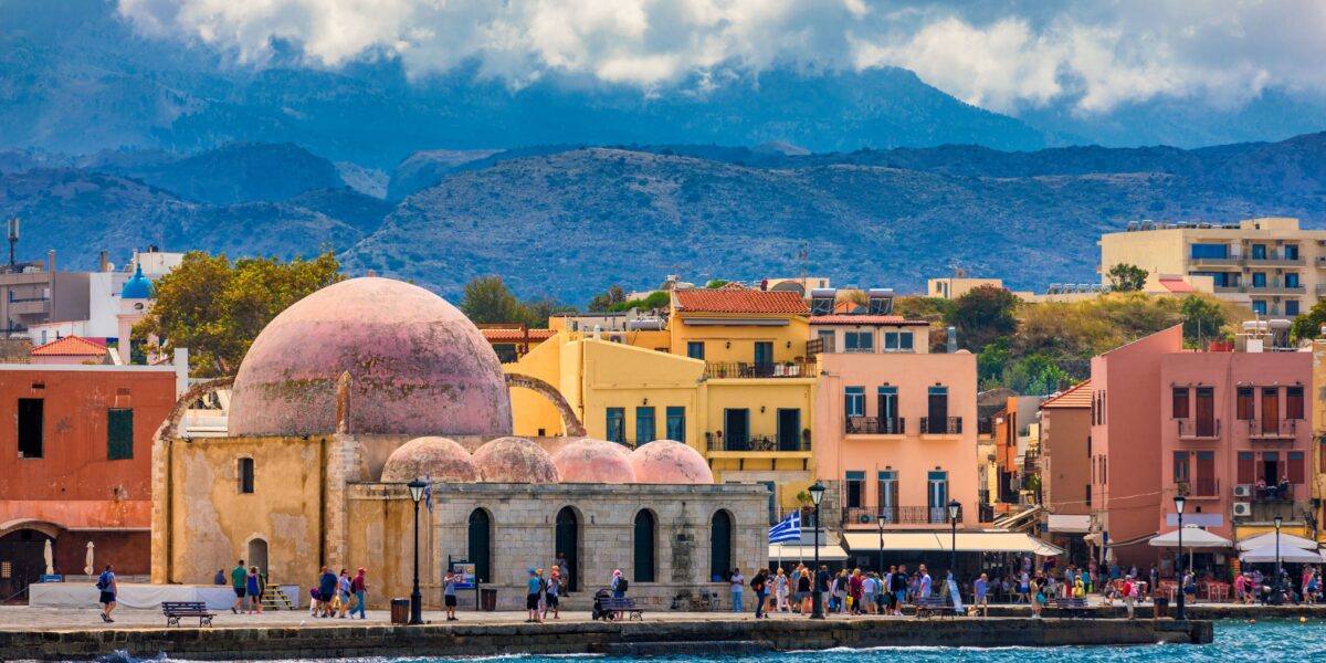 A scenic view of Chania in Crete featuring colorful buildings, a historic domed structure, and people walking along the waterfront, with a backdrop of mountainous terrain and a partly cloudy sky.