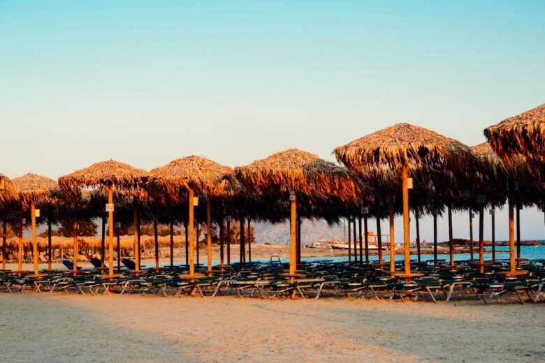 Rows of straw parasols and empty sunbeds on the sandy shore of Elafonisi Beach in Crete, with the sea and distant mountains visible in the background during sunset.