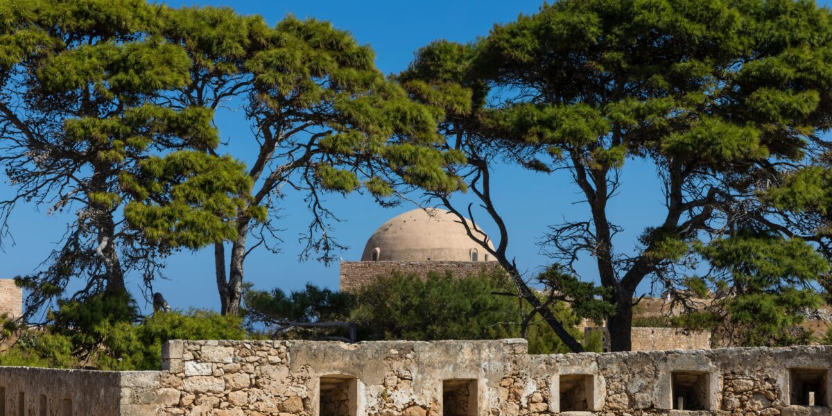 A view of Rethymnon, showing an old stone wall in the foreground with green trees partially obscuring a large, domed building in the background under a clear blue sky.