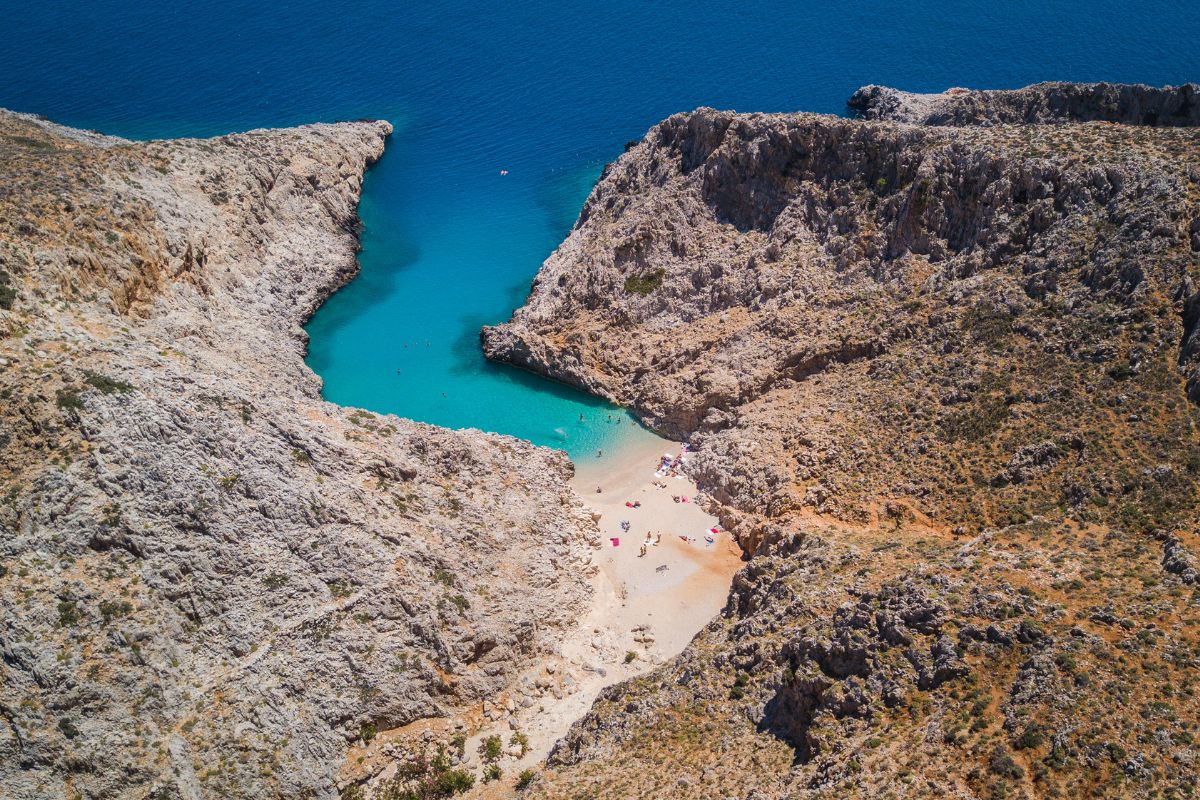 Aerial view of Seitan Limani beach with clear turquoise waters, surrounded by rocky cliffs and sparse vegetation. People are sunbathing and swimming at the shore.