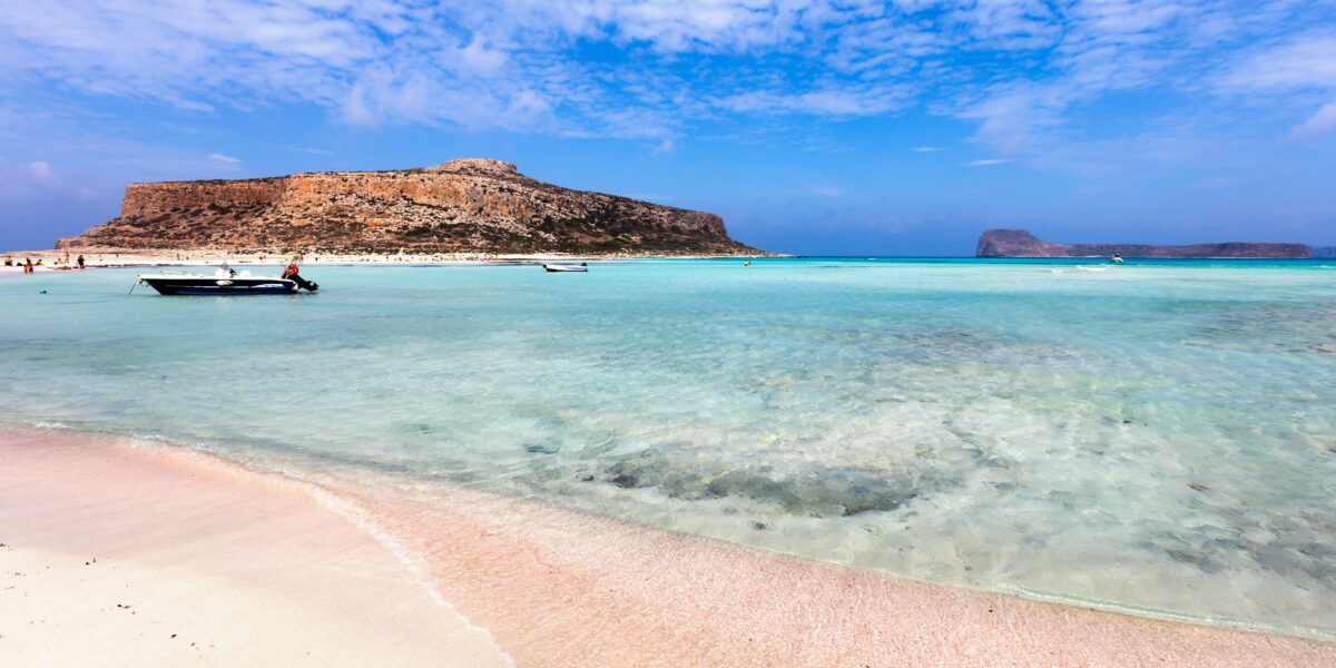 A scenic view of Balos Beach featuring clear turquoise waters, a sandy shoreline, and a rugged, rocky hill in the background. A few small boats are anchored near the shore, and people can be seen enjoying the beach. The sky is partly cloudy with a blue hue.