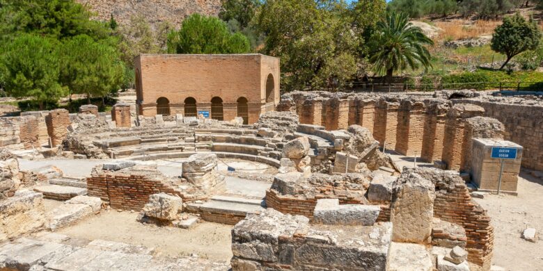 The image shows the ruins of the Odeon at Gortyna, featuring ancient stone and brick structures, with a circular seating area surrounded by remnants of walls and columns. A sign indicating "Odeon" is visible, and the site is set against a backdrop of trees and hills.