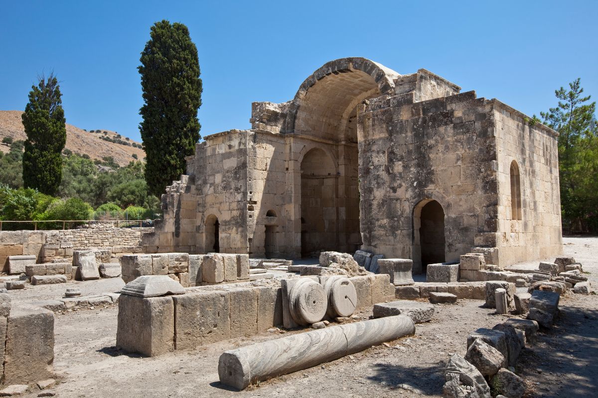 The image shows the ruins of a large stone structure at Gortyna, featuring arched doorways and remnants of columns and stones scattered around the site. Tall cypress trees and hills are visible in the background under a clear blue sky.