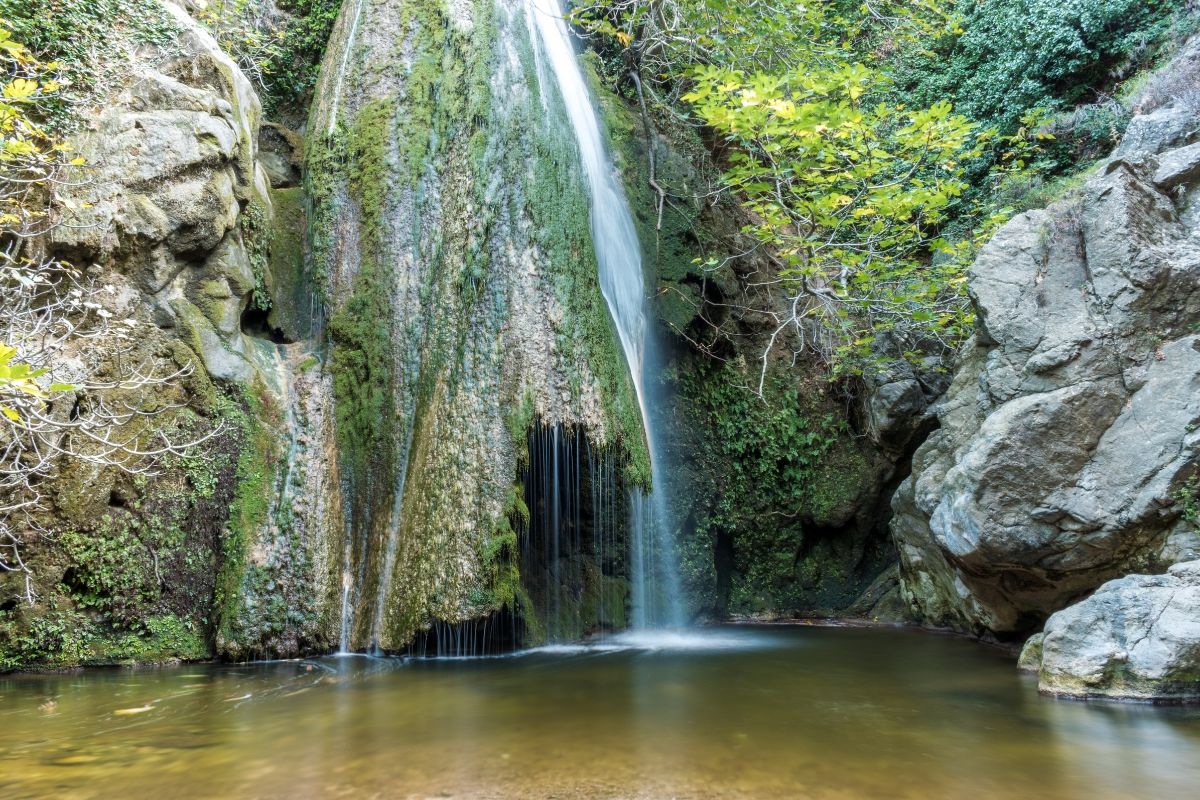 The image shows Richtis Waterfall in East Crete, with water cascading down a moss-covered rock face into a serene pool below, surrounded by lush greenery and rocky formations.