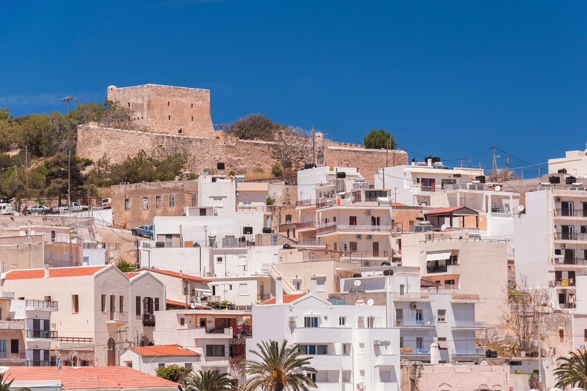 The image shows Sitia Castle situated on a hilltop, overlooking a town with white and beige buildings under a clear blue sky.