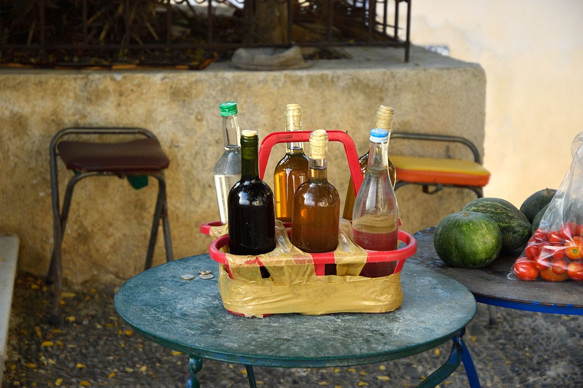 The image shows a small table with a red basket containing bottles of olive oil and wine. There are also some watermelons and a bag of tomatoes on the table.