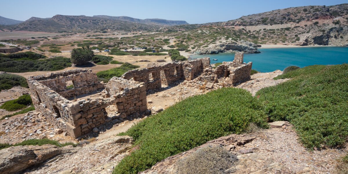 The image shows the ancient ruins of Itanos in East Crete, with stone walls and structures overlooking a scenic bay with turquoise water, surrounded by rugged hills and sparse vegetation.