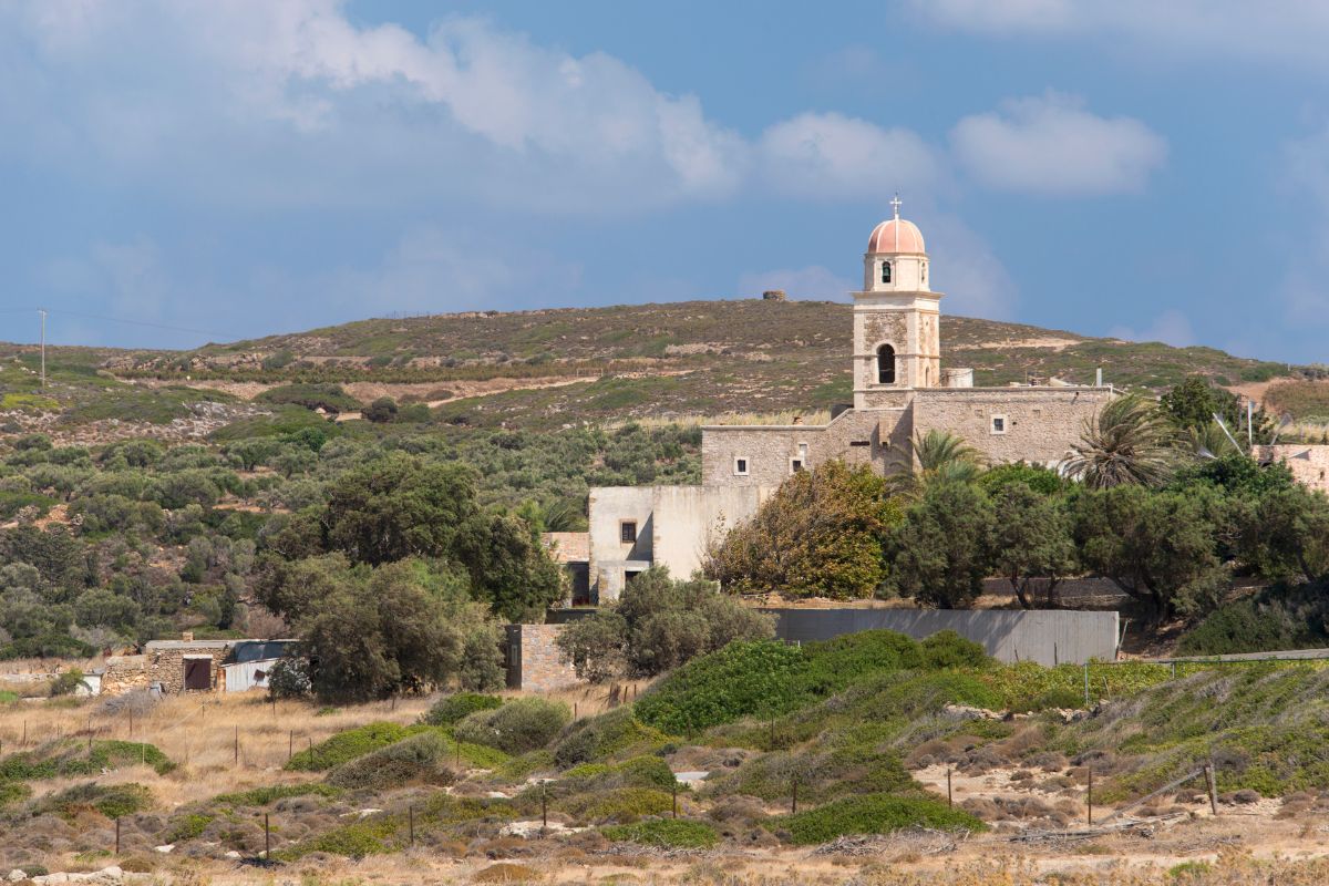 The image shows Toplou Monastery in Sitia, surrounded by greenery and set against a backdrop of hills under a partly cloudy sky.